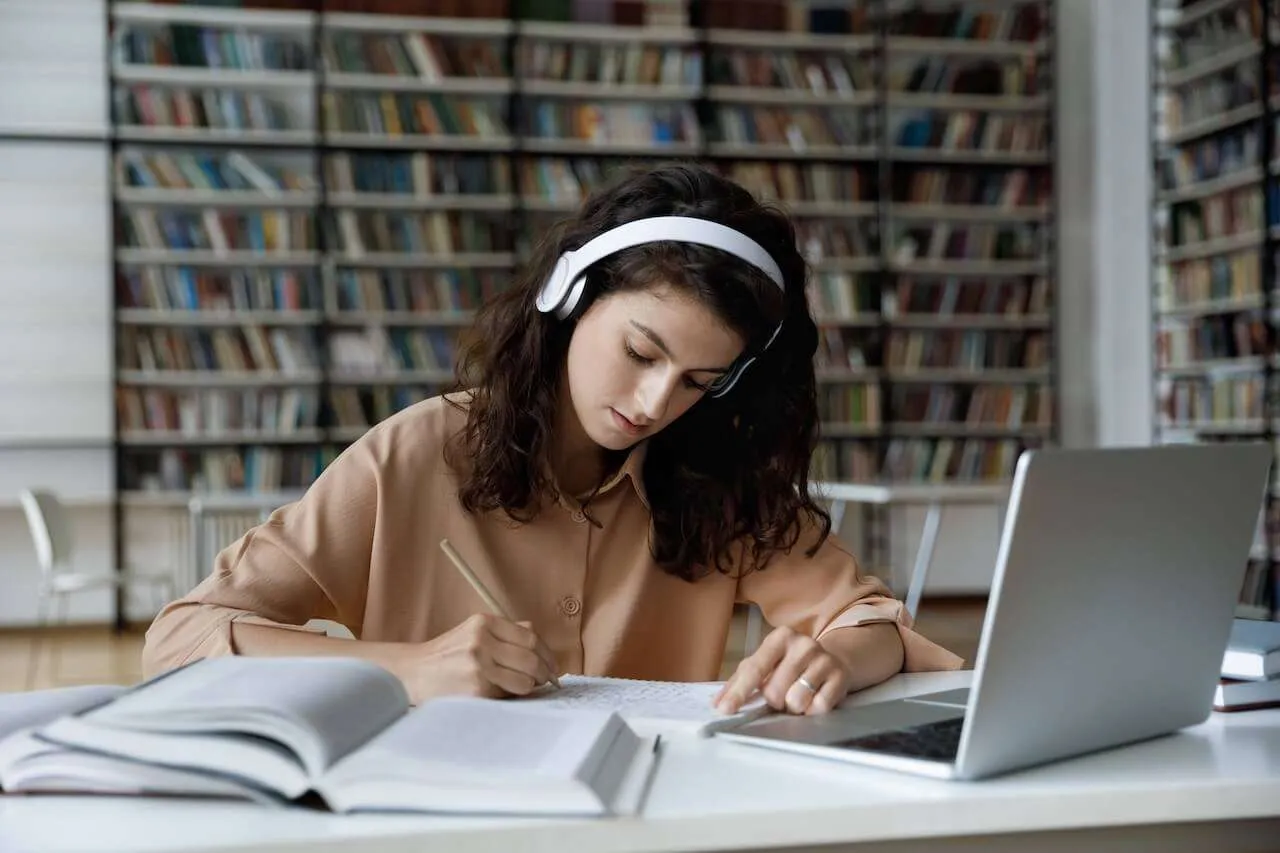 Picture of a woman wearing headphones writing at a desk in front of an open laptop.