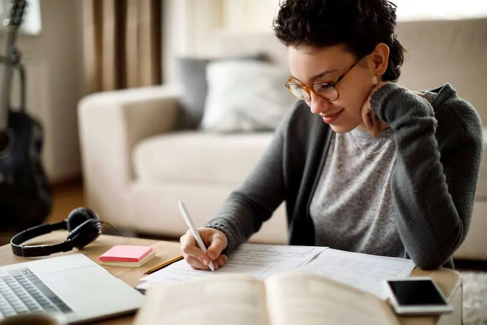 Image of a woman writing in a notebook on a desk covered with a laptop and papers