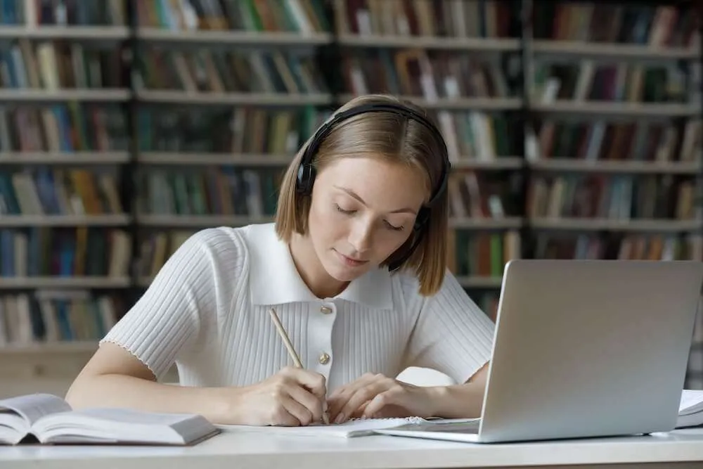 Woman watching a laptop whilst writing in a notebook.