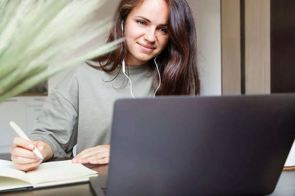Woman watching a laptop whilst writing in a notebook.