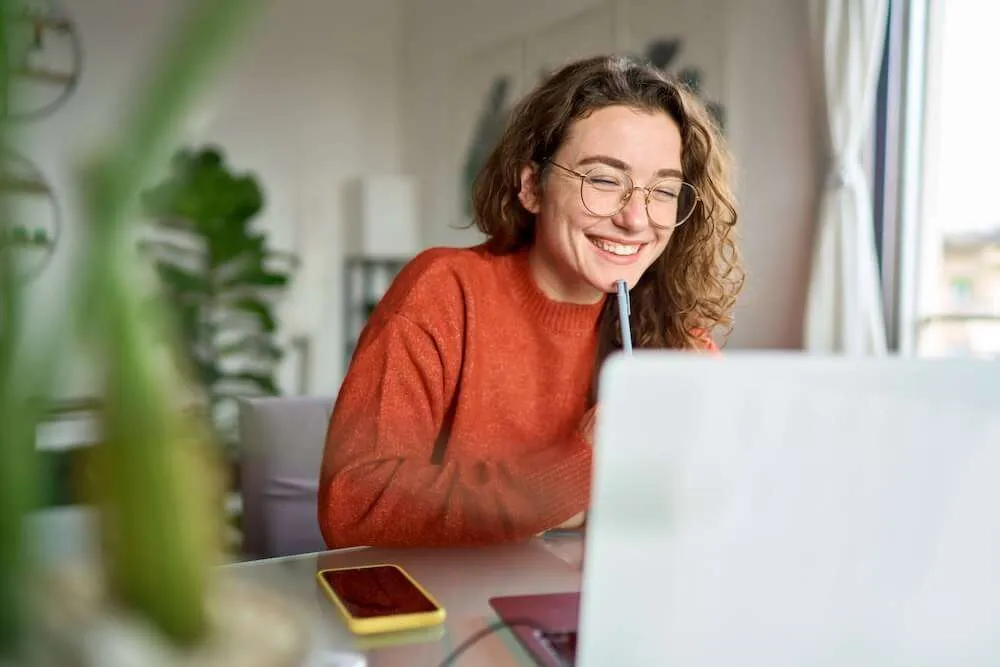 Image of a woman in an orange jumper smiling in front of a laptop.