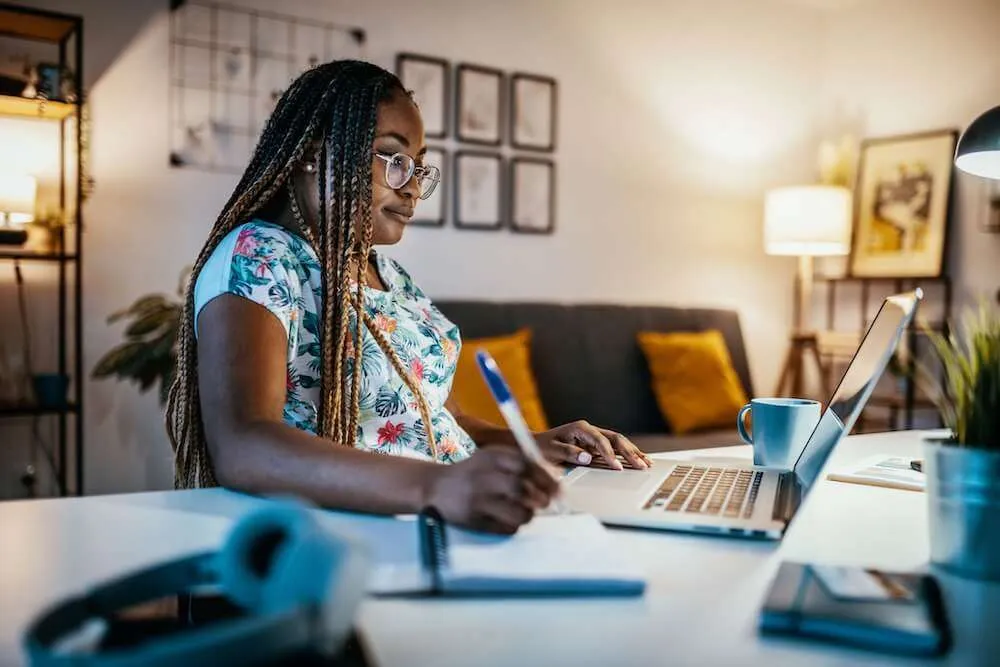 Woman watching a laptop whilst writing in a notebook.