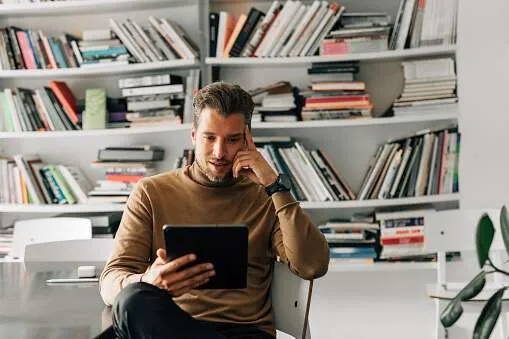 Photo of a man sitting in a chair with crossed legs reading something on an iPad.